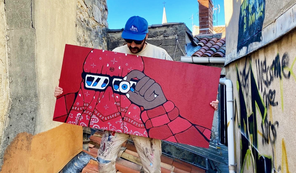 Artist Jeune Woof holding a red artwork of a figure with 3D glasses, standing on a rooftop with graffiti and a clear blue sky in the background.
