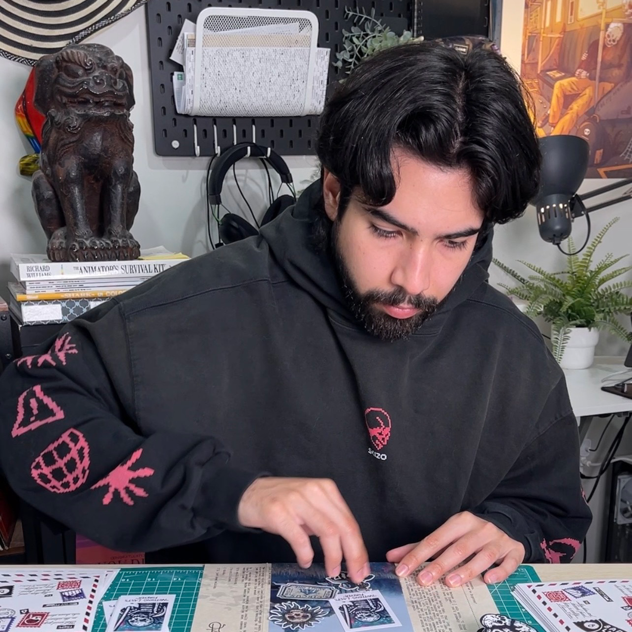 A man in a black hoodie sitting in his studio arranging stickers on the table in front of him, with sculptures and art in the background.