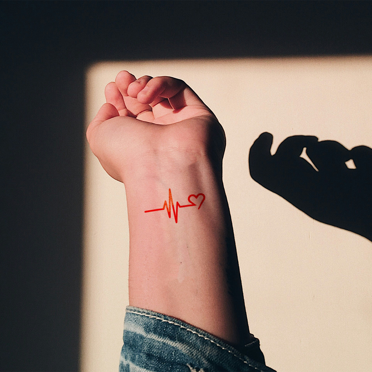 Close-up of a wrist with a red temporary tattoo of a heartbeat line ending in a small heart, lit by warm sunlight.