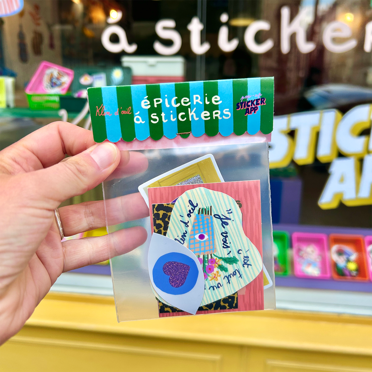 Hand holding a colorful packet of assorted stickers in front of a shop window displaying more sticker designs.