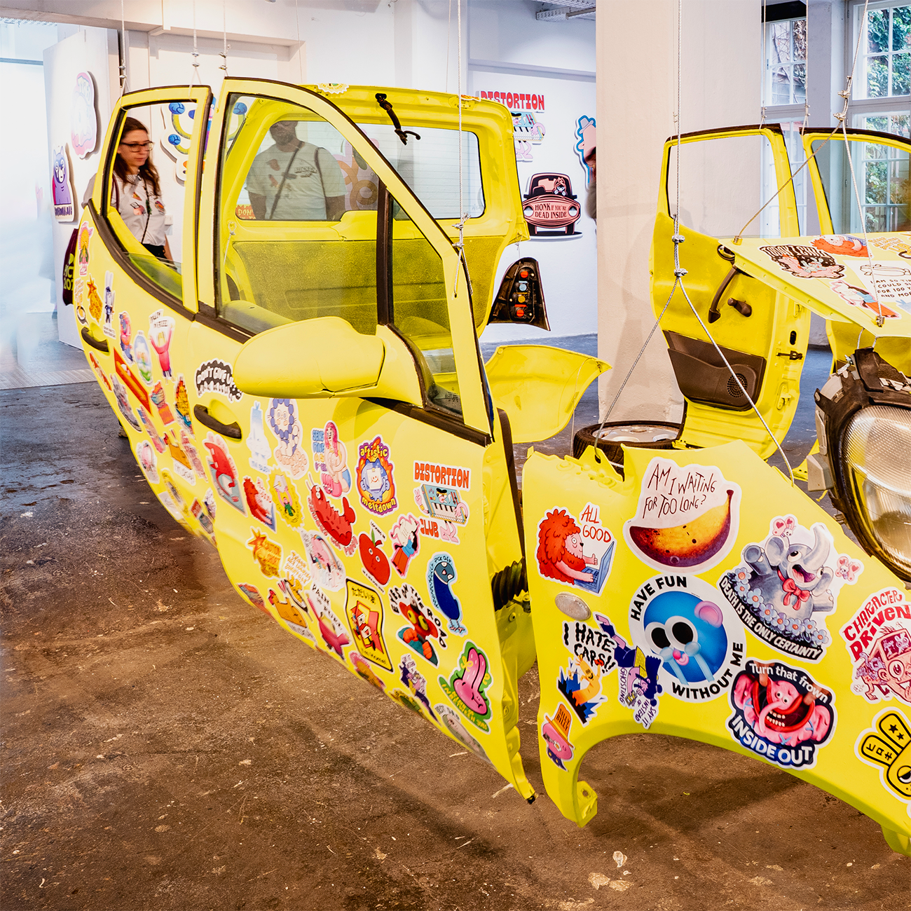 Yellow Mercedes body hanging in wires, covered in bumper stickers in various shapes and sizes at an art show.