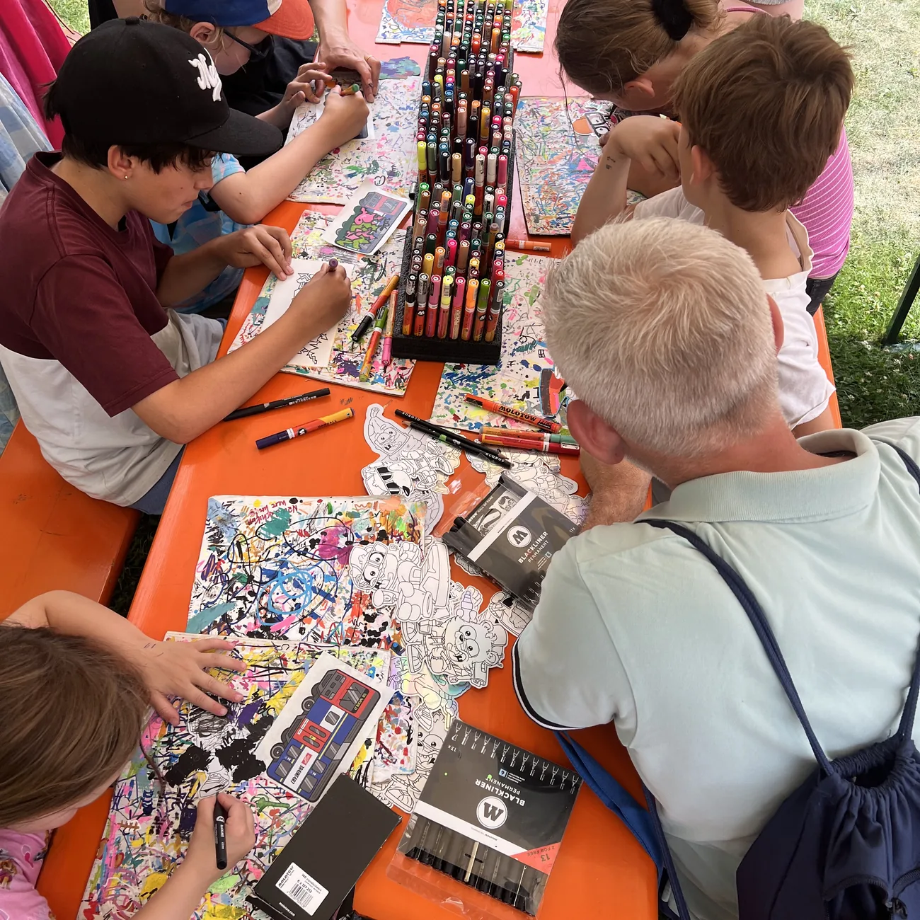 Kids coloring Brainfart die-cut stickers around a rack of paint markers at an outdoor festival workshop