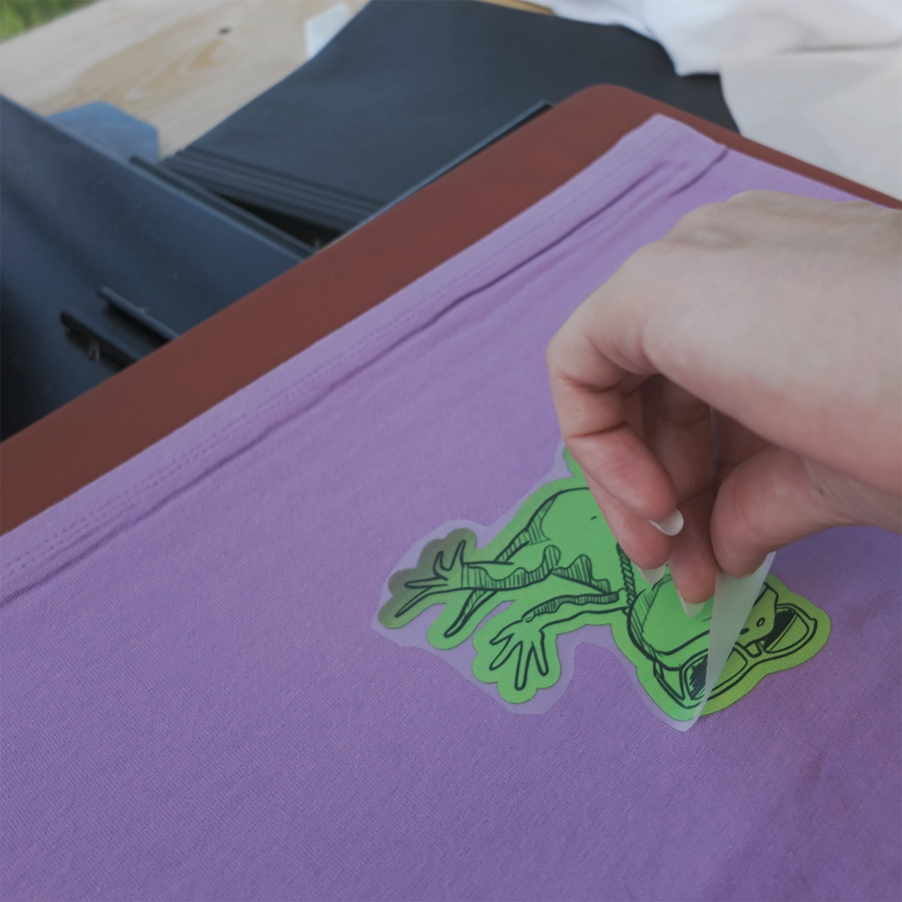 Die-cut DTF heat transfer print of a dark and light green frog placed on a purple garment with a heat press