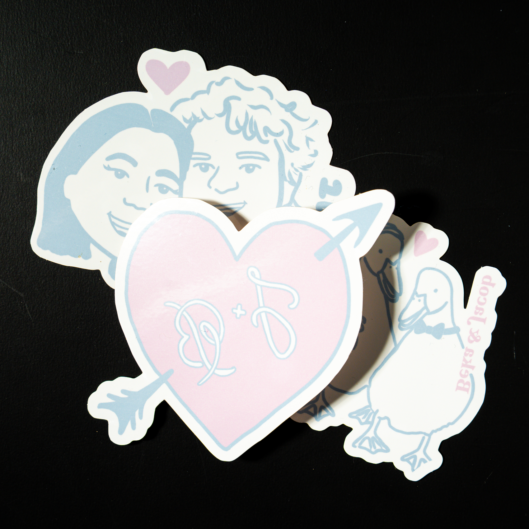 Pastel temporary tattoos featuring a heart with initials and arrows, a smiling illustrated couple, and a duck design with small pink hearts, arranged on a dark surface.
