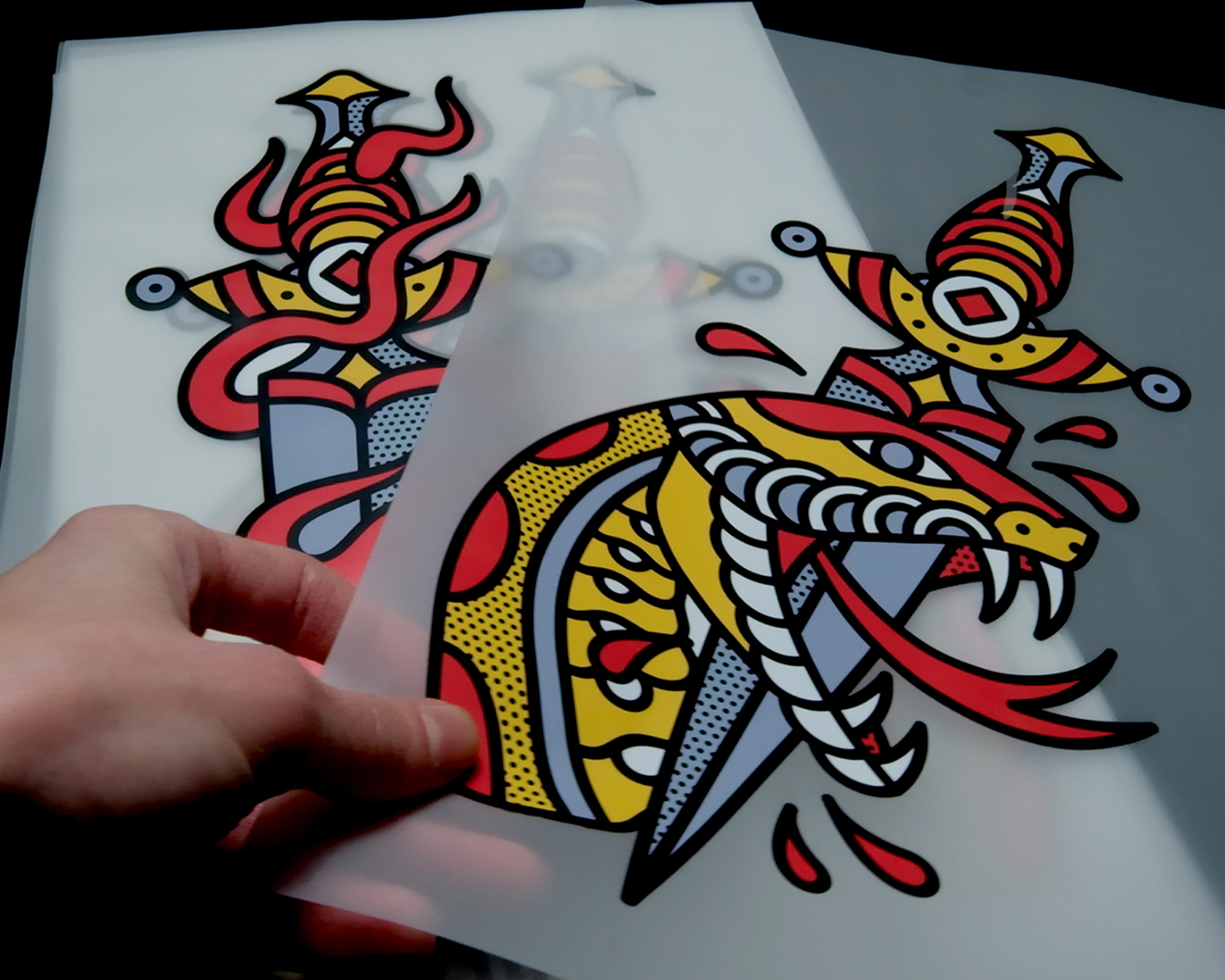 A hand holds a colorful transparent sheet with a cartoon-style snake and sword design in vibrant red, yellow, and black colors.