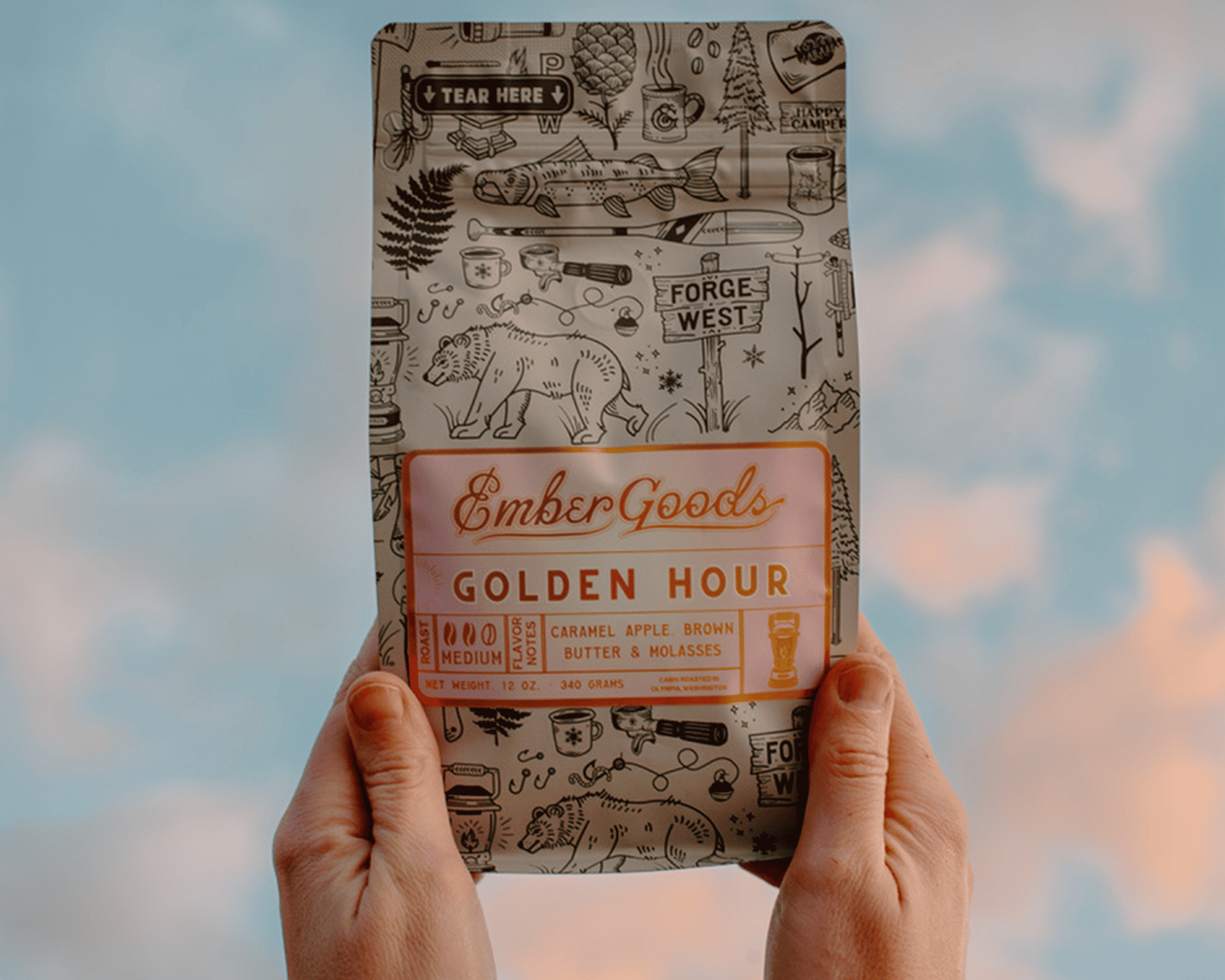 Hands holding a bag of Ember Goods Golden Hour coffee with a metallic label against a sky backdrop