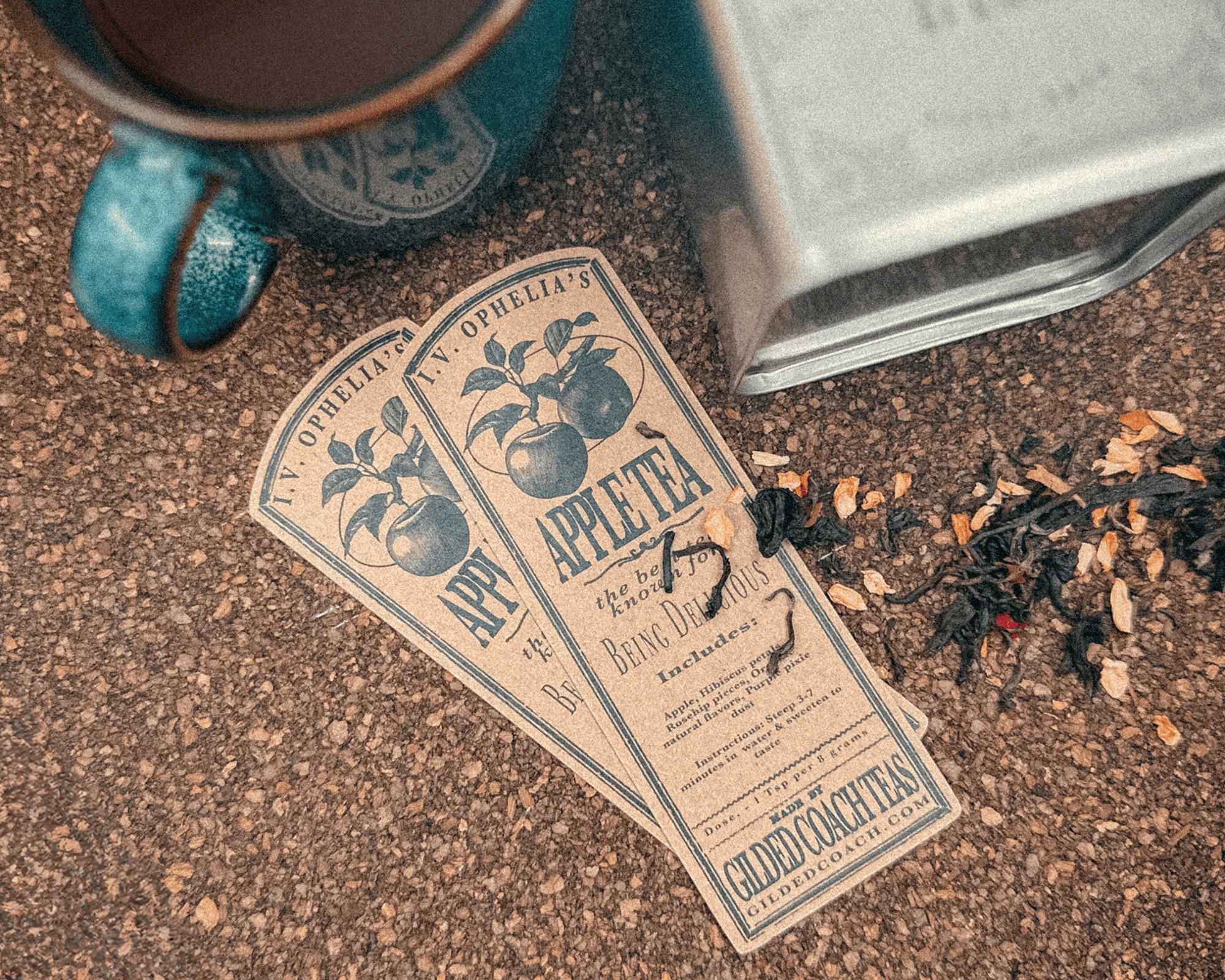 Two vintage-style tea kraft stickers with apple illustrations lie on a textured surface, next to a blue ceramic mug and scattered tea leaves.