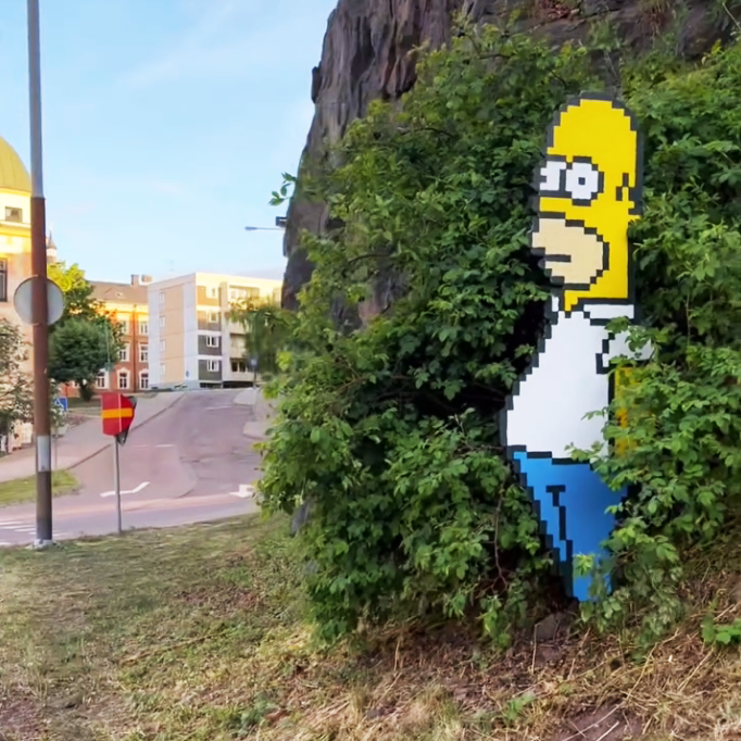 Large-scale Homer Simpson vinyl sticker peaking out of a bush in an urban environment.